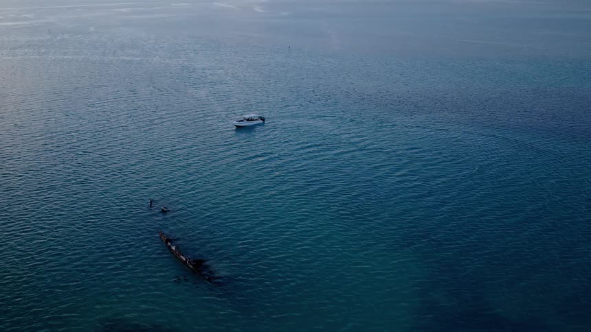 High aerial pull-back shot of a white motorboat anchored above a sunken shipwreck in the tranquil blue ocean as the sun reflects on the water