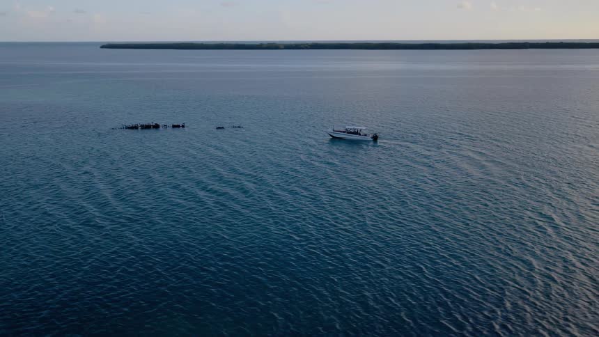 An aerial view pulls away from a speedboat and a sunken shipwreck in the tranquil open ocean as the sun begins to set