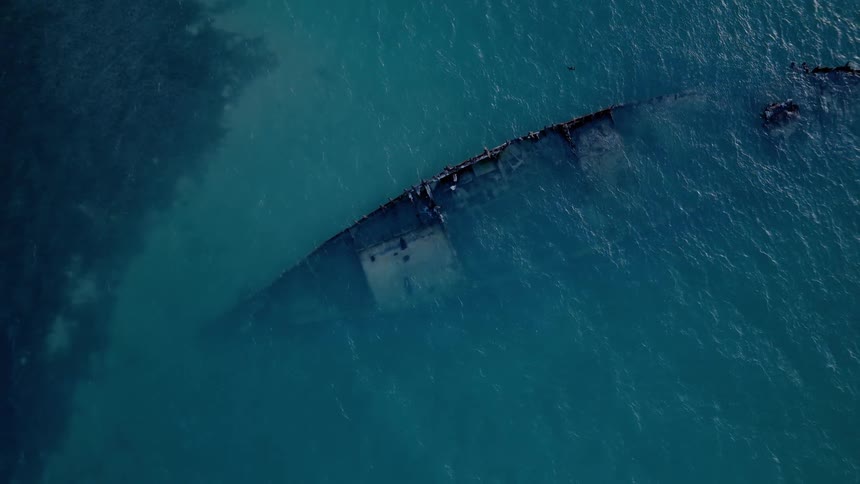 A haunting top-down aerial view of a shipwreck submerged just below the surface of the calm, turquoise ocean