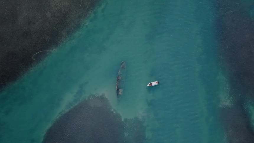 An aerial top-down view slowly descends towards a sunken shipwreck and a small boat in the clear, shallow turquoise sea