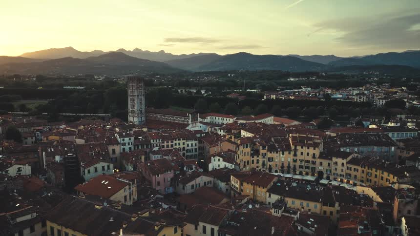 Seagulls fly over the historic cityscape of Lucca, Italy, as the sun sets behind the distant mountains