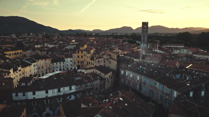 A static aerial view captures the historic city of Lucca, Italy, with its terracotta rooftops and Piazza dell'Anfiteatro, bathed in the warm light of sunset