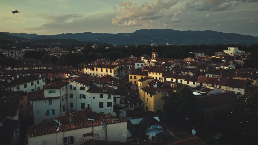 A scenic view over the historic rooftops of an Italian city at sunset, with mountains in the background and birds flying by