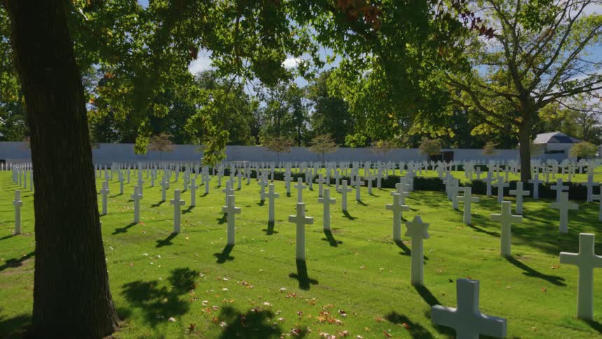 Rows of white crosses at a military cemetery stand peacefully on a sunny day under the shade of trees