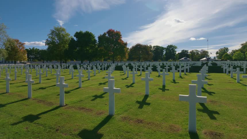 Rows of white crosses stand in a military cemetery on a bright sunny day, casting long shadows across the green grass
