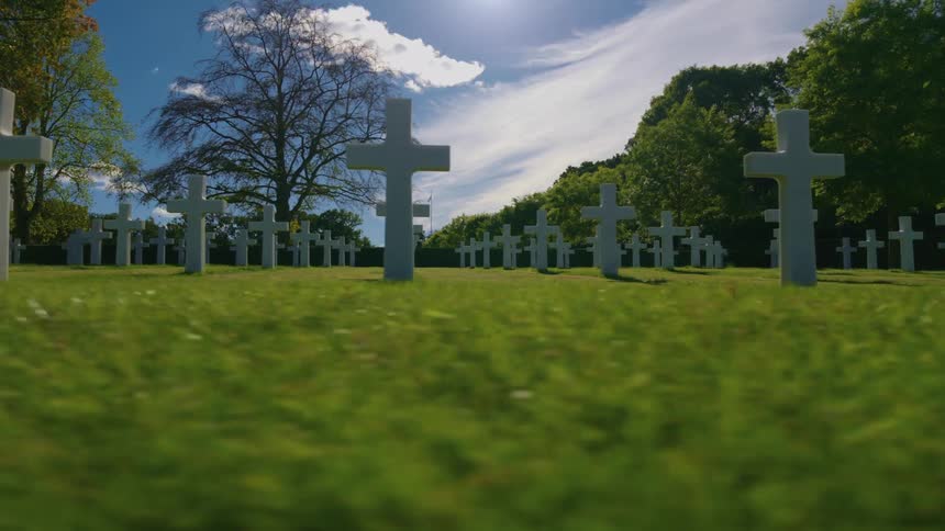 A slow, low-angle tracking shot reveals rows of white crosses in a serene, sunlit military cemetery