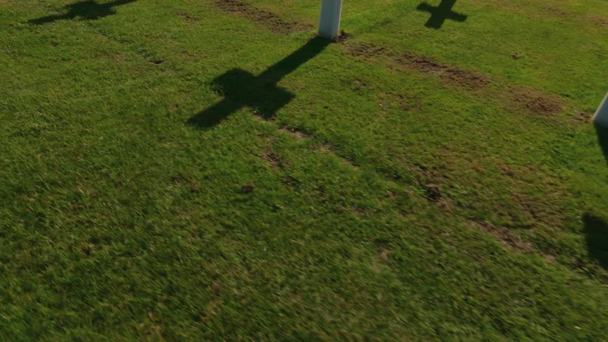 Shadows of white cross grave markers are cast on the lush green grass of a memorial cemetery