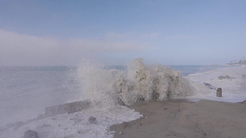 Powerful ocean waves crash in dramatic slow motion against a dilapidated concrete seawall on a sandy beach