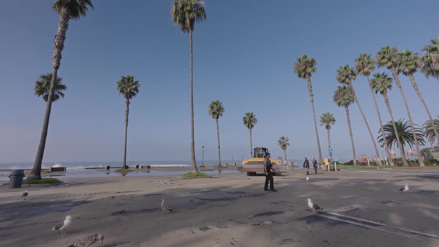 A man walks past a bulldozer on a flooded beachside road as cleanup begins after high tide