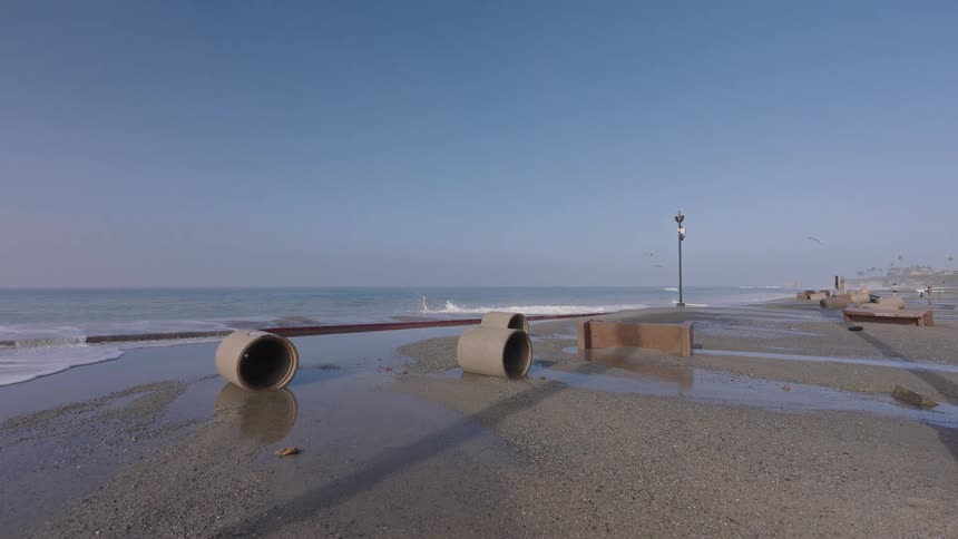 A time-lapse of gentle ocean waves washing ashore on a sandy beach scattered with construction debris and concrete pipes under a clear blue sky