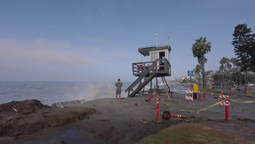 Spectators watch in slow motion as a massive ocean wave crashes against the shoreline next to a lifeguard tower