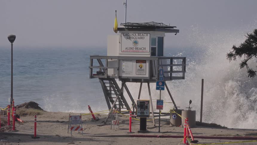 A massive ocean wave crashes ashore in slow motion next to a lifeguard tower at a closed-off Aliso Beach in Laguna Beach