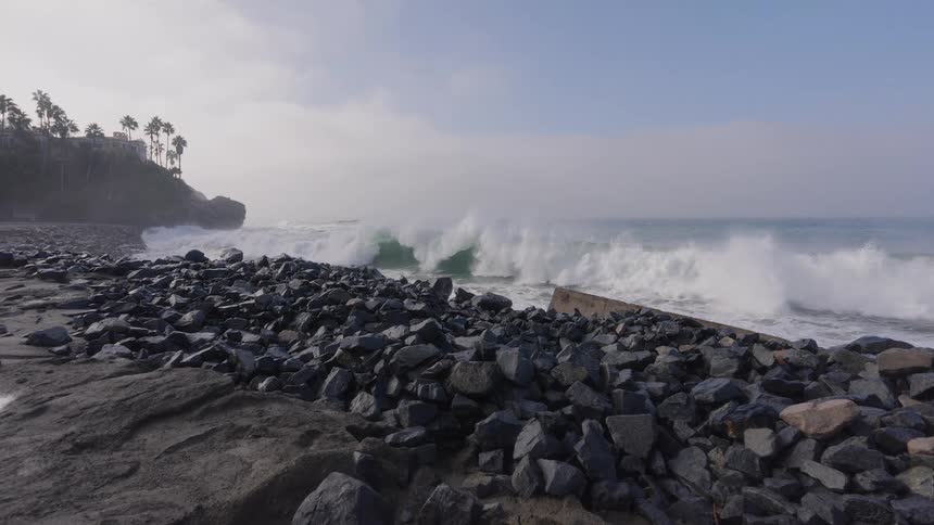 A massive ocean wave crashes in slow motion towards a rocky shoreline on a foggy day