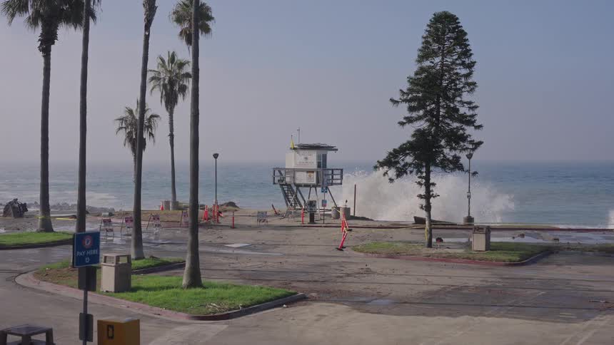 A powerful wave crashes over a seawall in slow motion during a high surf event, flooding an empty beach parking lot with a lifeguard tower in the background