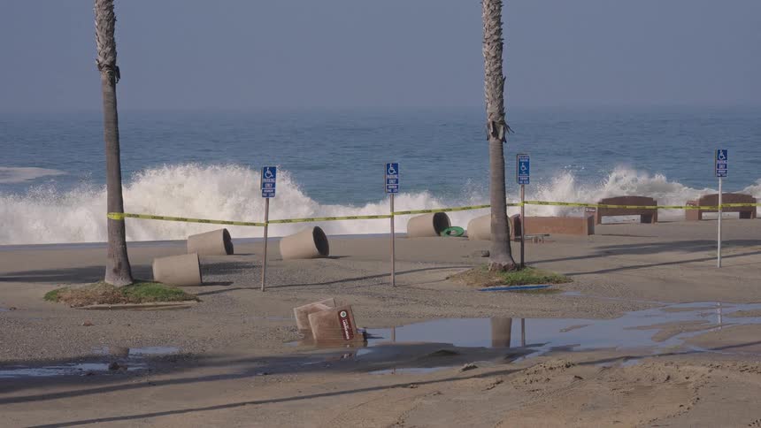 A beach parking lot is flooded and cordoned off with caution tape as powerful storm waves crash ashore in slow motion