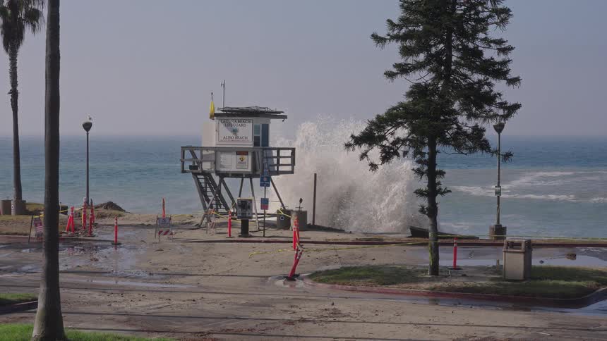 A powerful ocean wave crashes behind a lifeguard tower in slow motion during a coastal storm, flooding the nearby beach and road