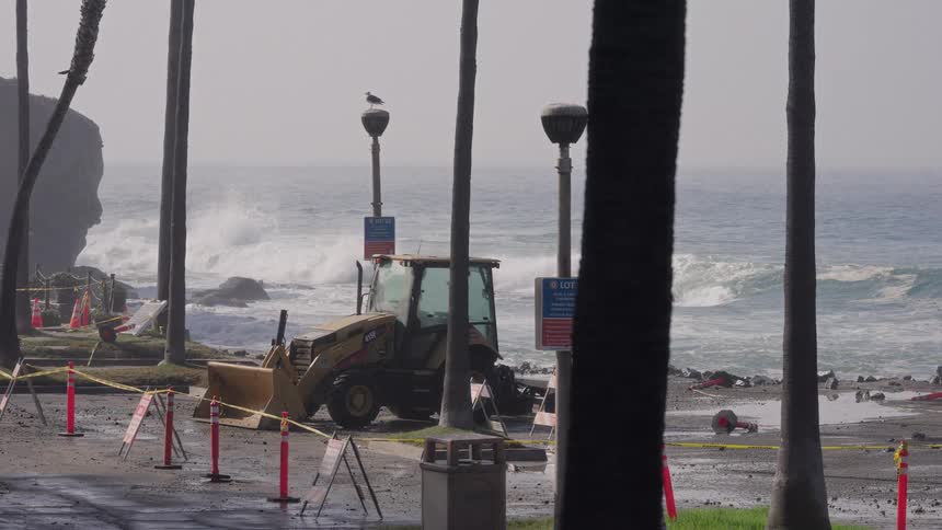 A slow-motion shot of a yellow backhoe on a closed coastal road with large ocean storm waves crashing in the background