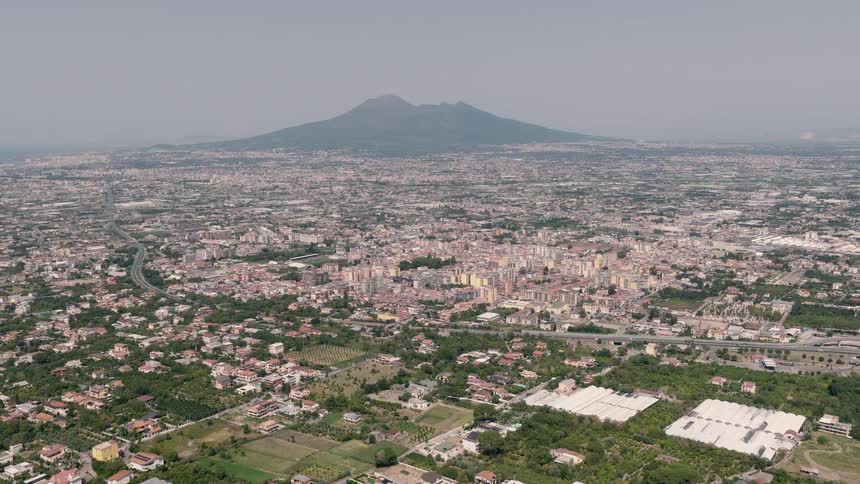 A slow aerial push-in shot over a sprawling city in Italy with the majestic Mount Vesuvius looming in the hazy background