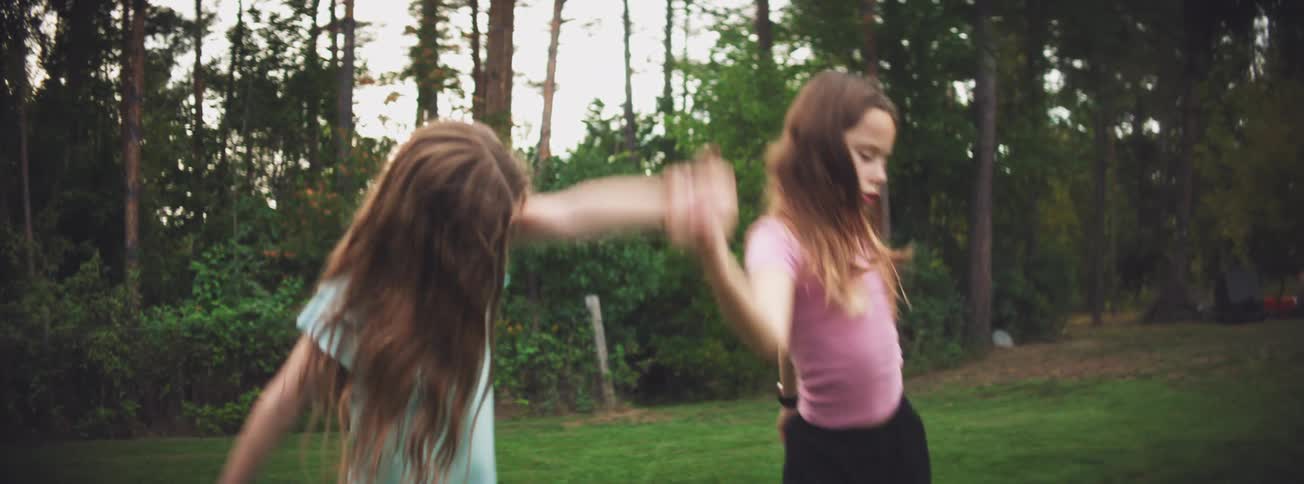 Two joyful young girls with long hair spin around in slow motion while holding hands on a grassy lawn
