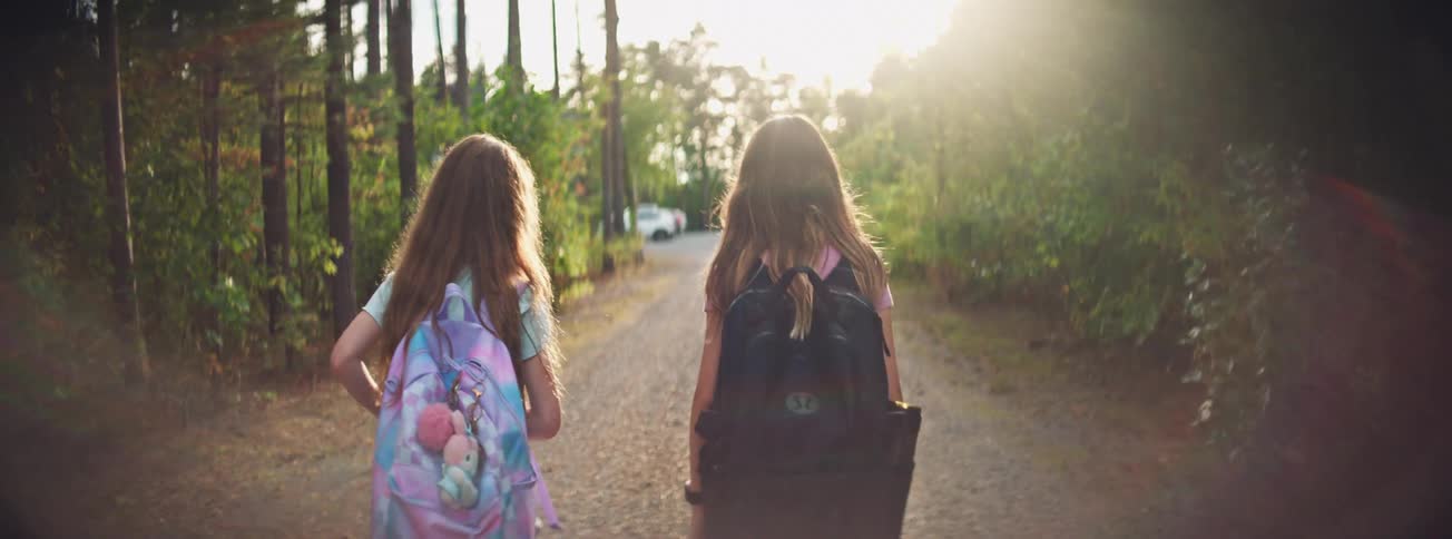 Two schoolgirls with backpacks walk down a rural path bathed in golden sunlight