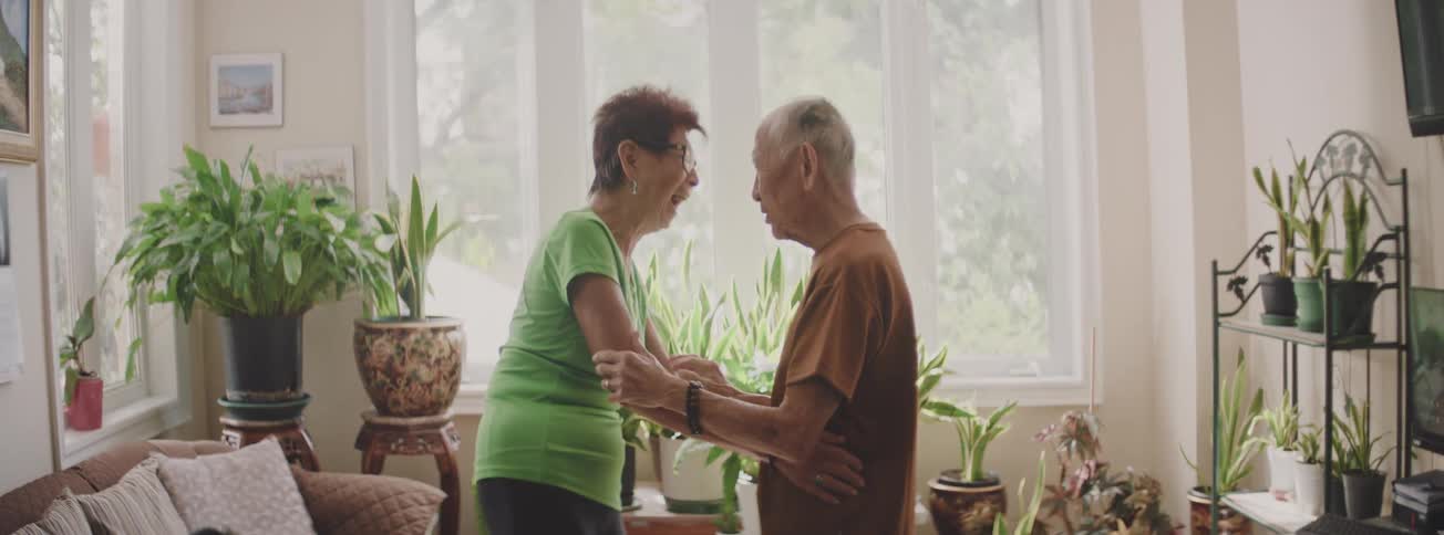 A joyful elderly Asian couple shares a laugh while dancing together in their bright, plant-filled living room