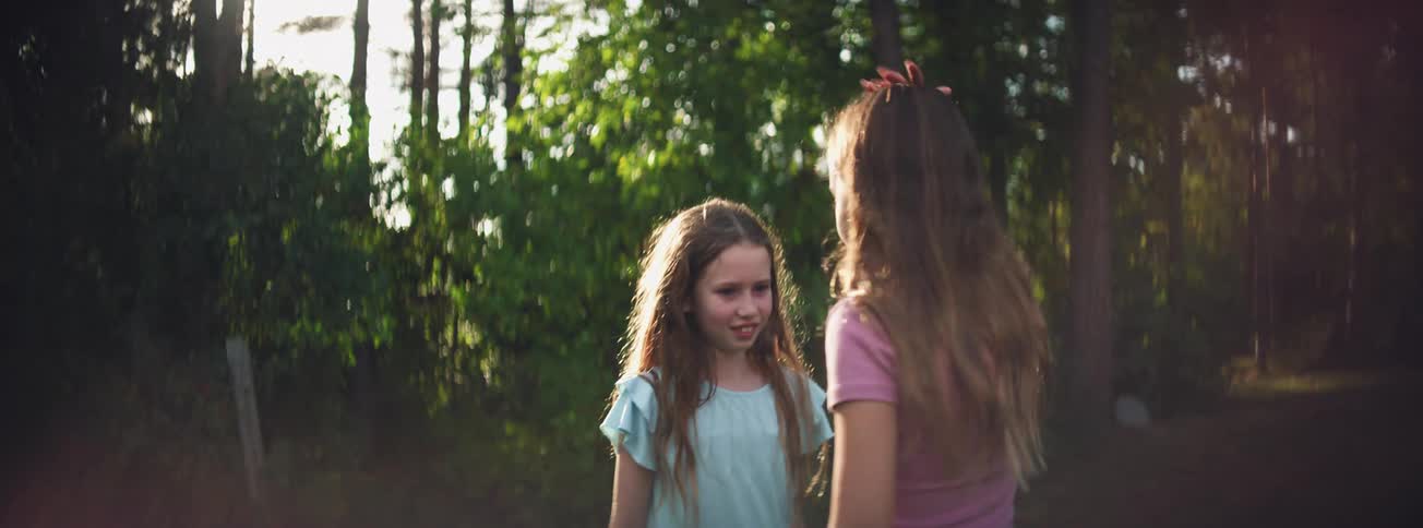 Two smiling young girls play a game outdoors, with one patting the other's head in a sunlit park before running off
