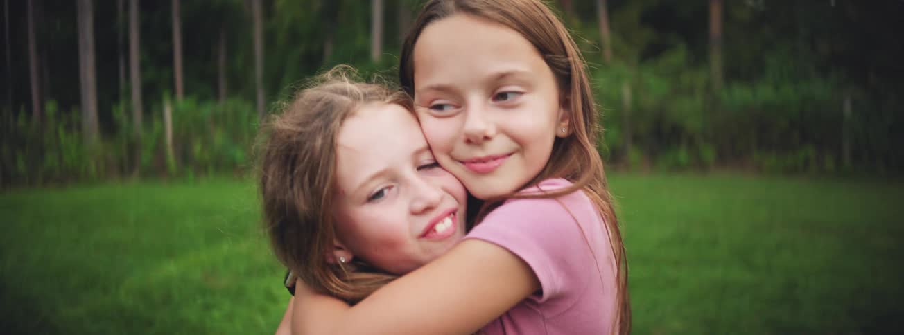 Two happy young sisters share a warm embrace and laugh while playing together outdoors in a grassy park