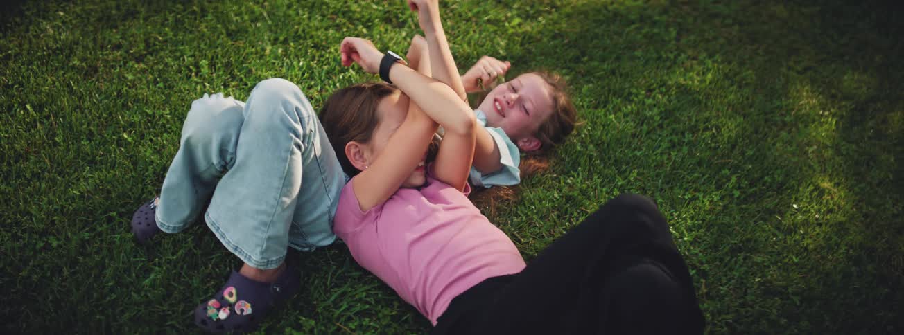 Two young sisters laugh and playfully wrestle while lying together on the lush green grass