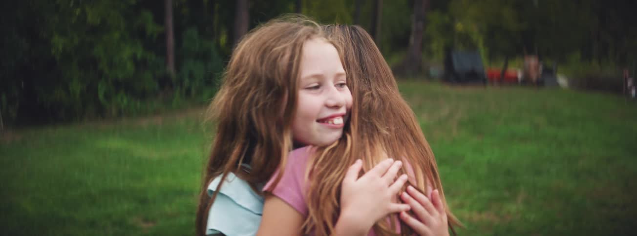 Two joyful young girls with long hair laugh and share a warm hug in slow motion while playing outside
