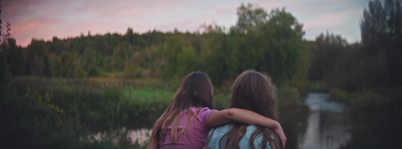 Two sisters embrace while sharing a peaceful moment watching the sunset over a tranquil river in the countryside