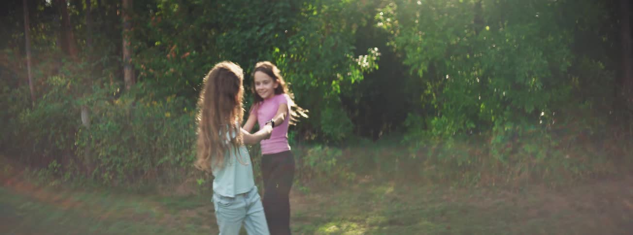 Two joyful young girls with long hair spin around holding hands in a sun-dappled park in slow motion