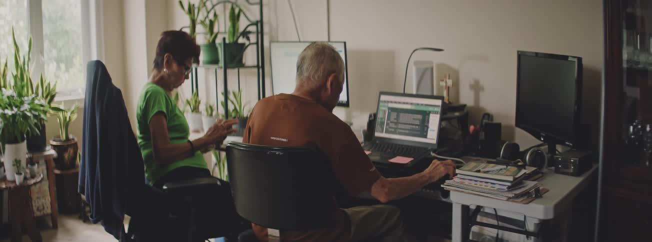 An elderly couple sits side-by-side in their home office, each working on their own computer