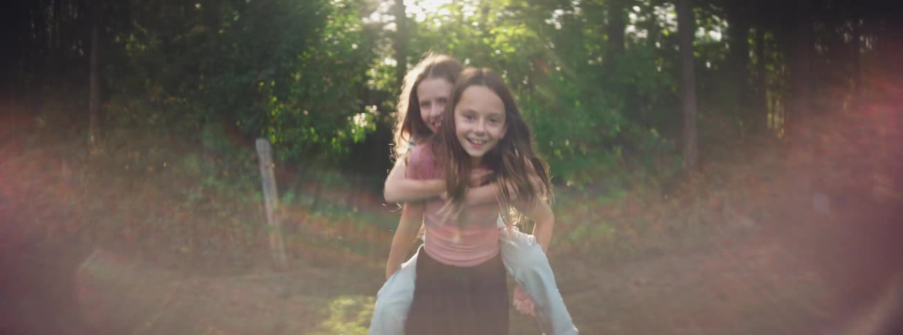Two joyful young sisters play in a sunlit field as one gives the other a piggyback ride in slow motion