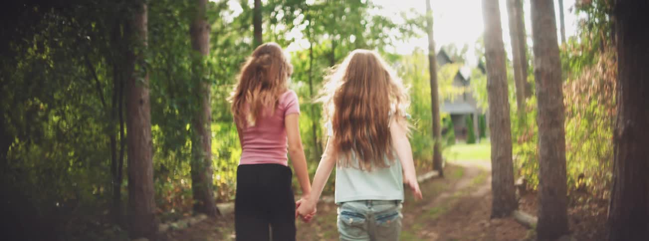 Two young girls hold hands and run together down a sun-drenched wooded path in slow motion