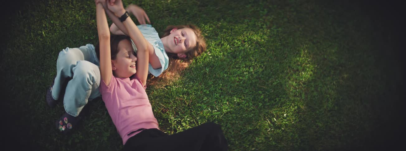 From a high angle, two carefree young girls laugh and playfully wrestle while relaxing together on a lush green lawn