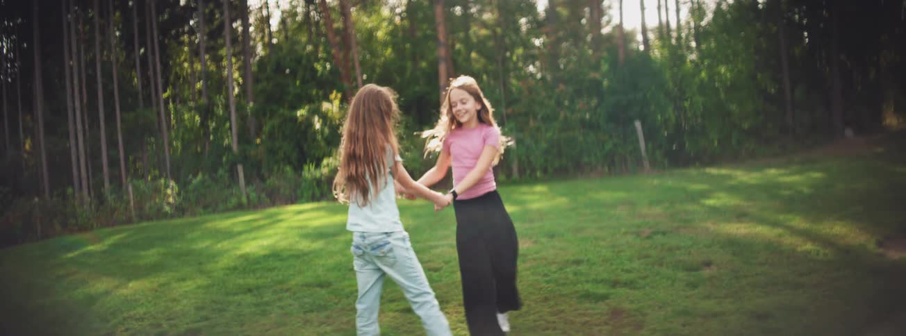 Two happy young girls with long hair spin and dance together in slow motion in a sunny green field