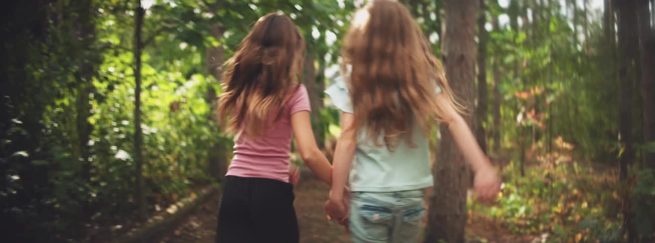 From behind, two young girls with long flowing hair hold hands while walking down a sunlit path in the woods in slow motion
