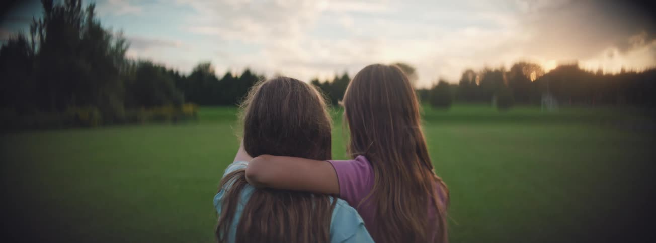 From behind, two young sisters stand together with their arms around each other in a green field, watching the sunset
