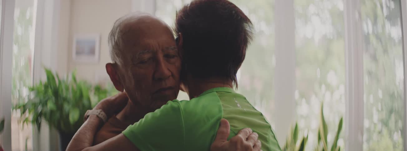 An elderly man with his eyes closed receives a comforting hug from a woman in an emotional embrace