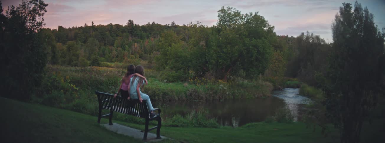 Two young girls sit together on a park bench at dusk, enjoying the tranquil view of a river and forest