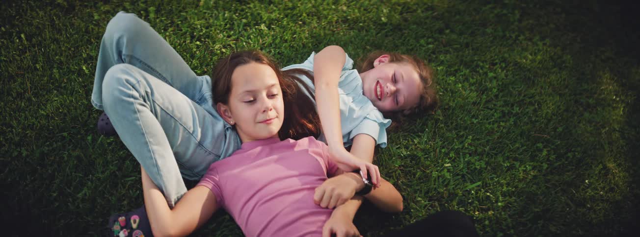 Two happy young sisters laugh and playfully tickle each other while lying on the green grass