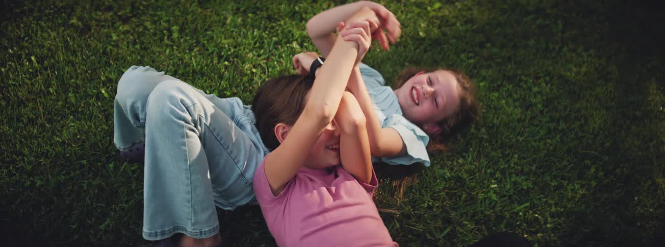 Two cheerful young girls laugh and playfully wrestle while lying together on the lush green grass on a sunny day