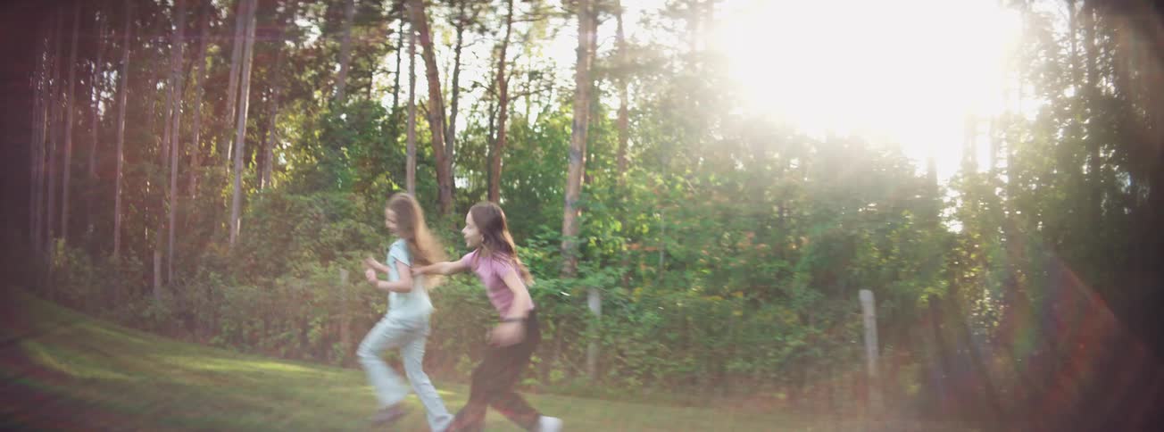 Two happy young girls playfully chase each other in a sunlit grassy field in slow motion