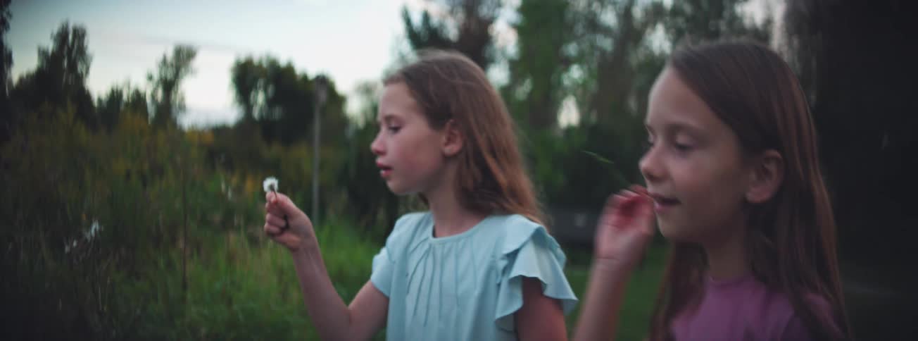 Two young sisters make a wish as they blow dandelion seeds in a park in slow motion