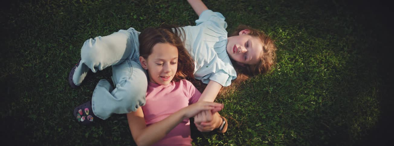 A top-down slow-motion view of two joyful young girls laughing and playing while lying on the green grass