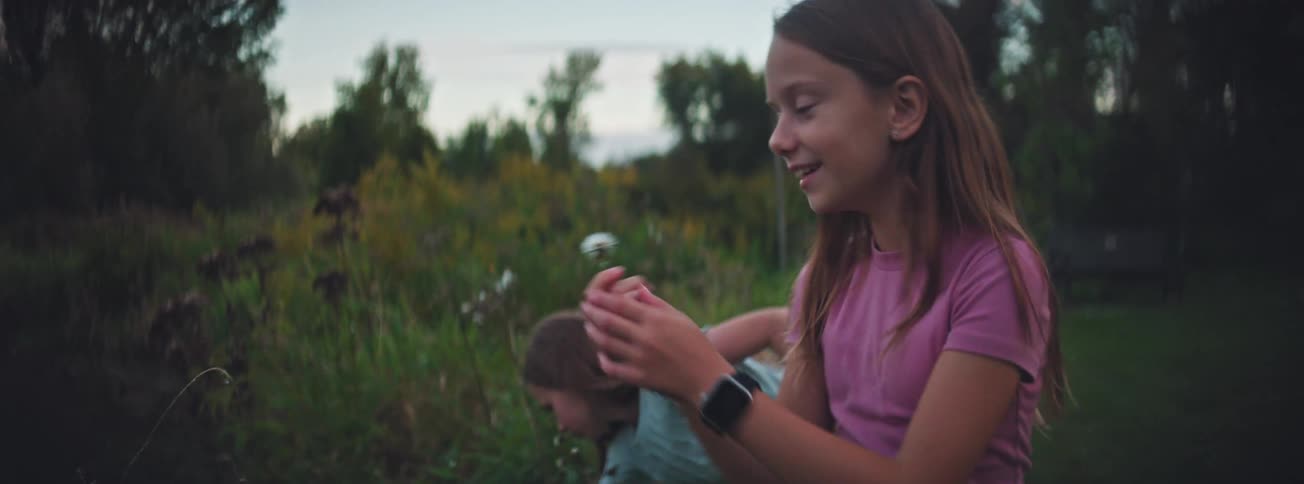 A young girl in a pink shirt blows on a dandelion, making a wish in a green field as her sister plays in the background