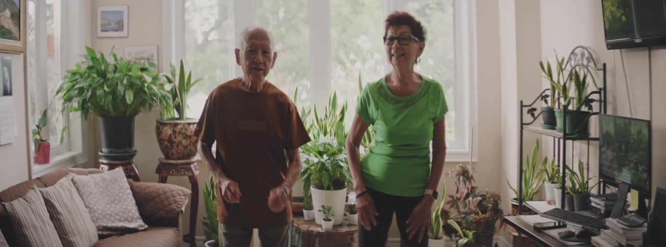 A happy and active senior couple exercises together at home in their living room surrounded by plants
