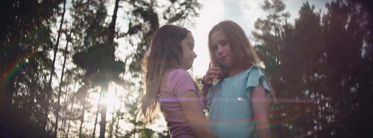 A young girl tenderly comforts her emotional friend as they stand together in a sun-flared forest