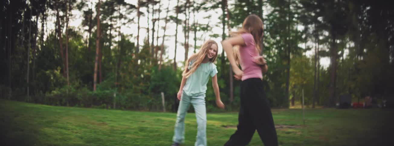 Two joyful young girls with long hair dance and play together in slow motion on a grassy lawn with a forest behind them