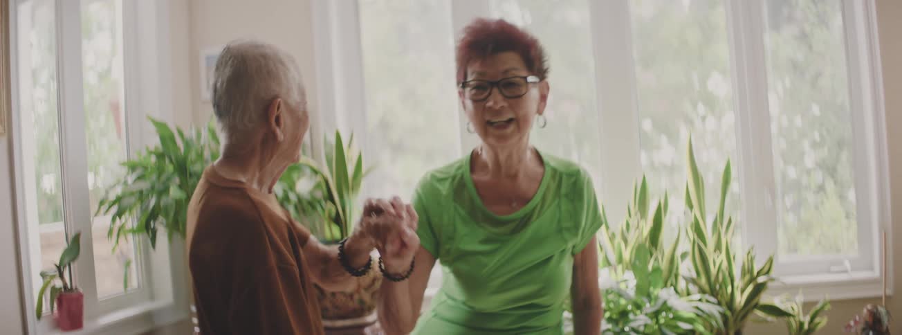 An elderly Asian couple shares a tender moment, laughing and holding each other while looking out the window of their plant-filled home